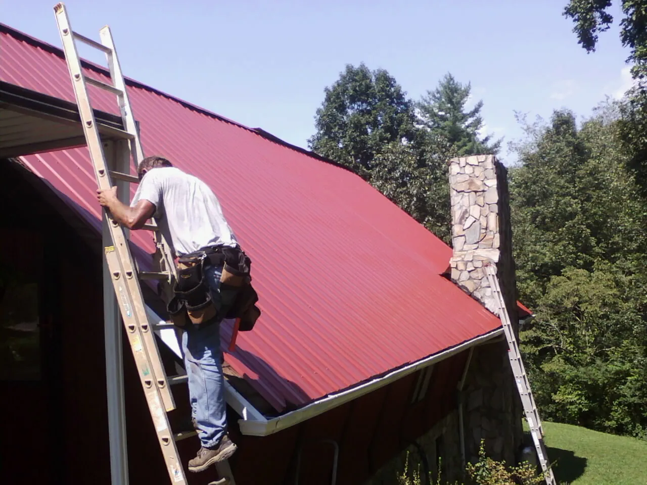 Roofer on a ladder beside a red metal roof and stone chimney — Soffit & Fascia Repair by Roofing Brothers in Parkville
